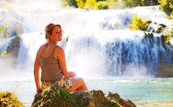 Erstaunliche junge Frau, die an einem Wasserfall Krka im Nationalpark, lächelnd reizend über ihrer Schulter sitzt