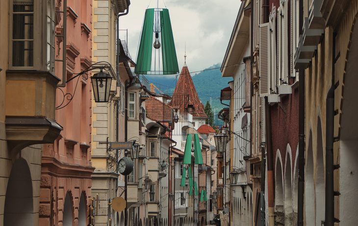 Blick durch die Häuserschlucht der Meraner Laubengasse mit ihren grünen, lampionartige Lampen - © joerghartmannphoto - stock.adobe.com