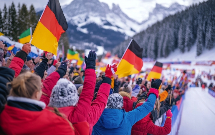 Begeisterte Biathlonfans in Winterjacken, die Deutschlandfahnen schwenken und die Athleten anfeuern, mit verschneiter Biathlonstrecke, Wald und Bergen im Hintergrund.
