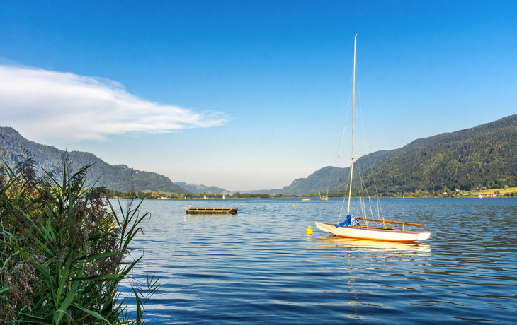 Segelboot auf einem ruhigen See mit bewaldeten Hügeln im Hintergrund und blauem Himmel. - ©zauberblicke - stock.adobe.com