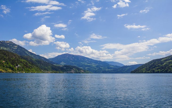 Segelboot auf einem ruhigen See mit bewaldeten Hügeln im Hintergrund und blauem Himmel. - ©zauberblicke - stock.adobe.com