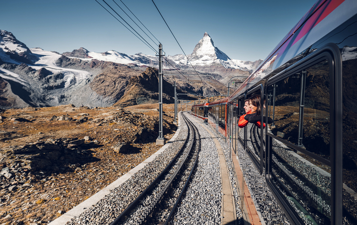 Schafe auf einer Wiese mit einem vorbeifahrenden rot-weißen Zug in einer Berglandschaft - © Gornergrat Bahn