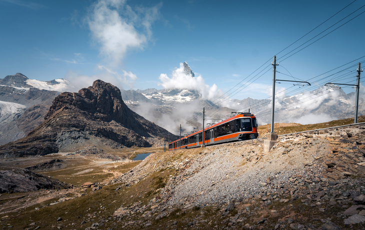 Schafe auf einer Wiese mit einem vorbeifahrenden rot-weißen Zug in einer Berglandschaft - © Gornergrat Bahn