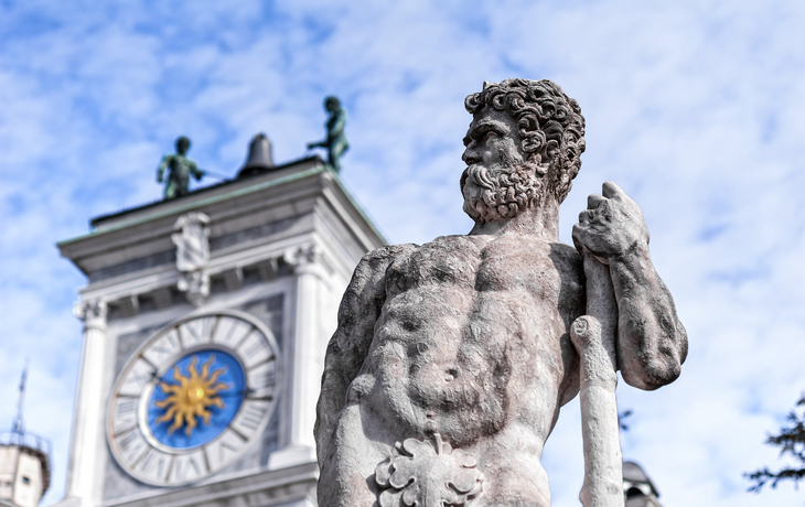Steinstatue eines bärtigen Mannes vor einem Gebäude mit Uhr und Figuren. - © Franco Nadalin