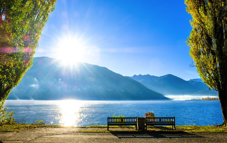 Sonnenaufgang über einem See mit Bergen im Hintergrund und Bäumen im Vordergrund. - ©fottoo - stock.adobe.com