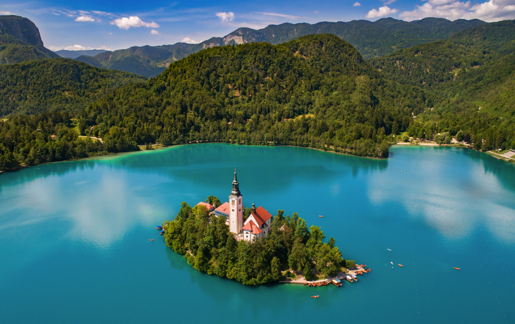 Insel mit Kirche in einem türkisfarbenen See, umgeben von bewaldeten Bergen. - © oneblink1 - stock.adobe.com